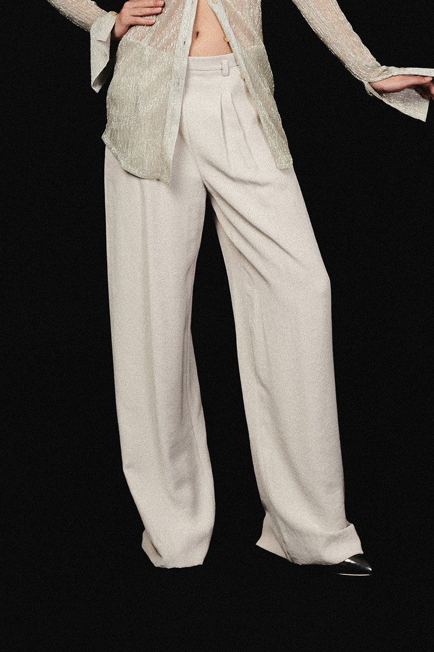 Woman in oyster wide leg, mid rise pants. Tailored waist front dart details, waist band with belt loops. Paired with sheer shimmer blouse.