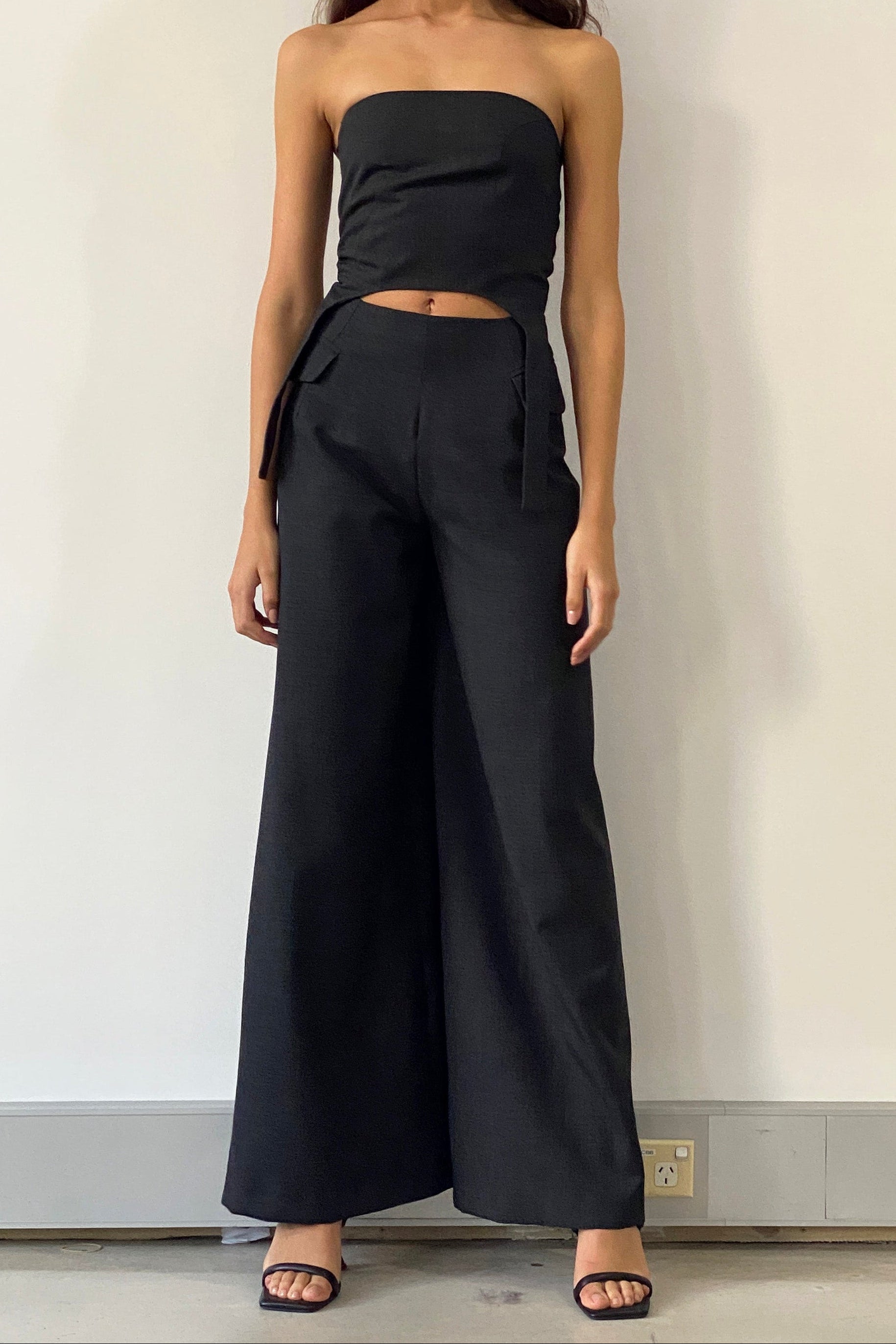Woman in black strapless top with curved hem and elongated strap details. Styled with black trousers.