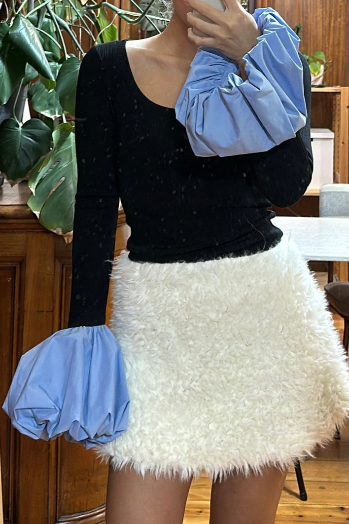 Woman in black long sleeve rib knit top with round neckline and blue balloon details on cuffs. Styled with a white fur mini skirt.
