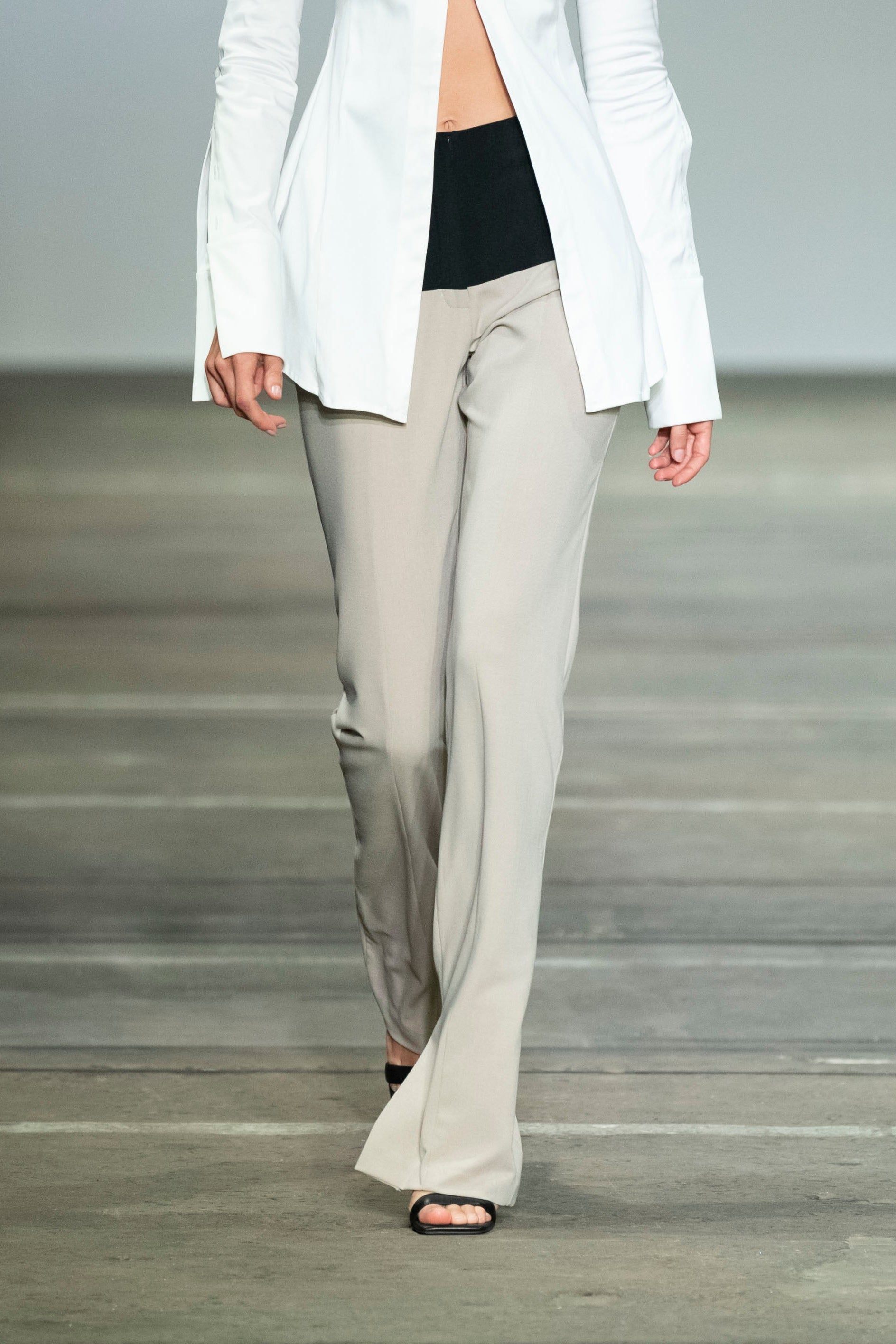 Woman in beige straight leg high waisted trousers. Contrast black waist panel with fly front and flap pocket details. Styled with white button up shirt.