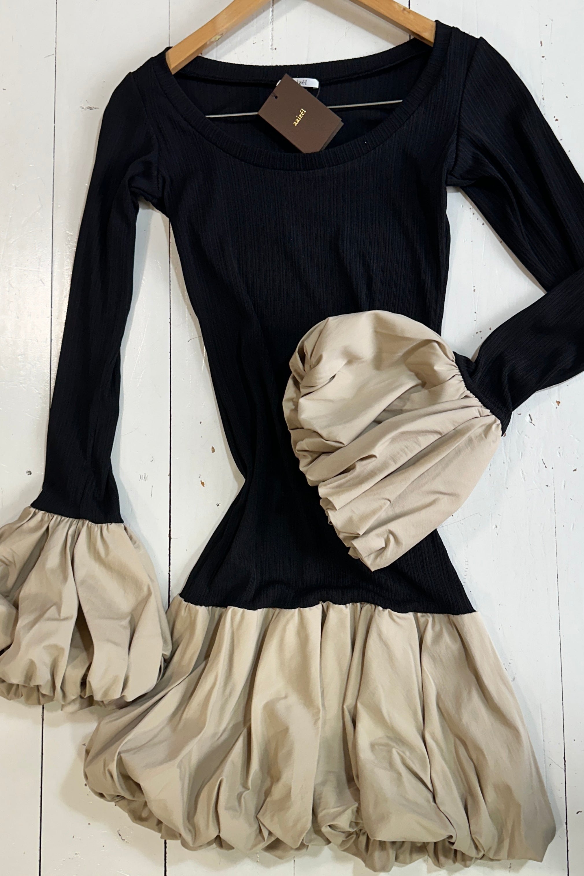 Flat lay of black mini dress with long sleeves and round neckline. Beige ballon details on sleeve cuffs and hem.