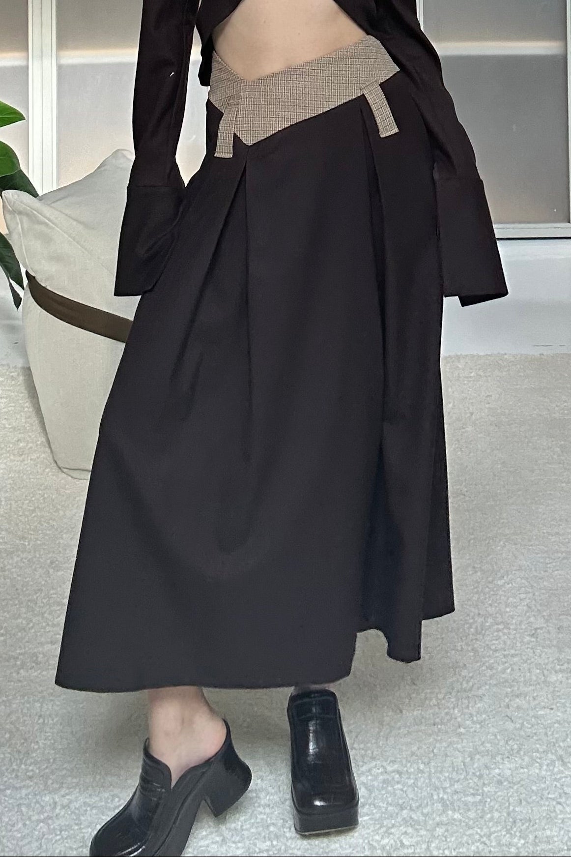 Woman in long skirt with box pleats and dipped waistband. Brown and beige check colourway. Styled with a dark brown cropped shirt.