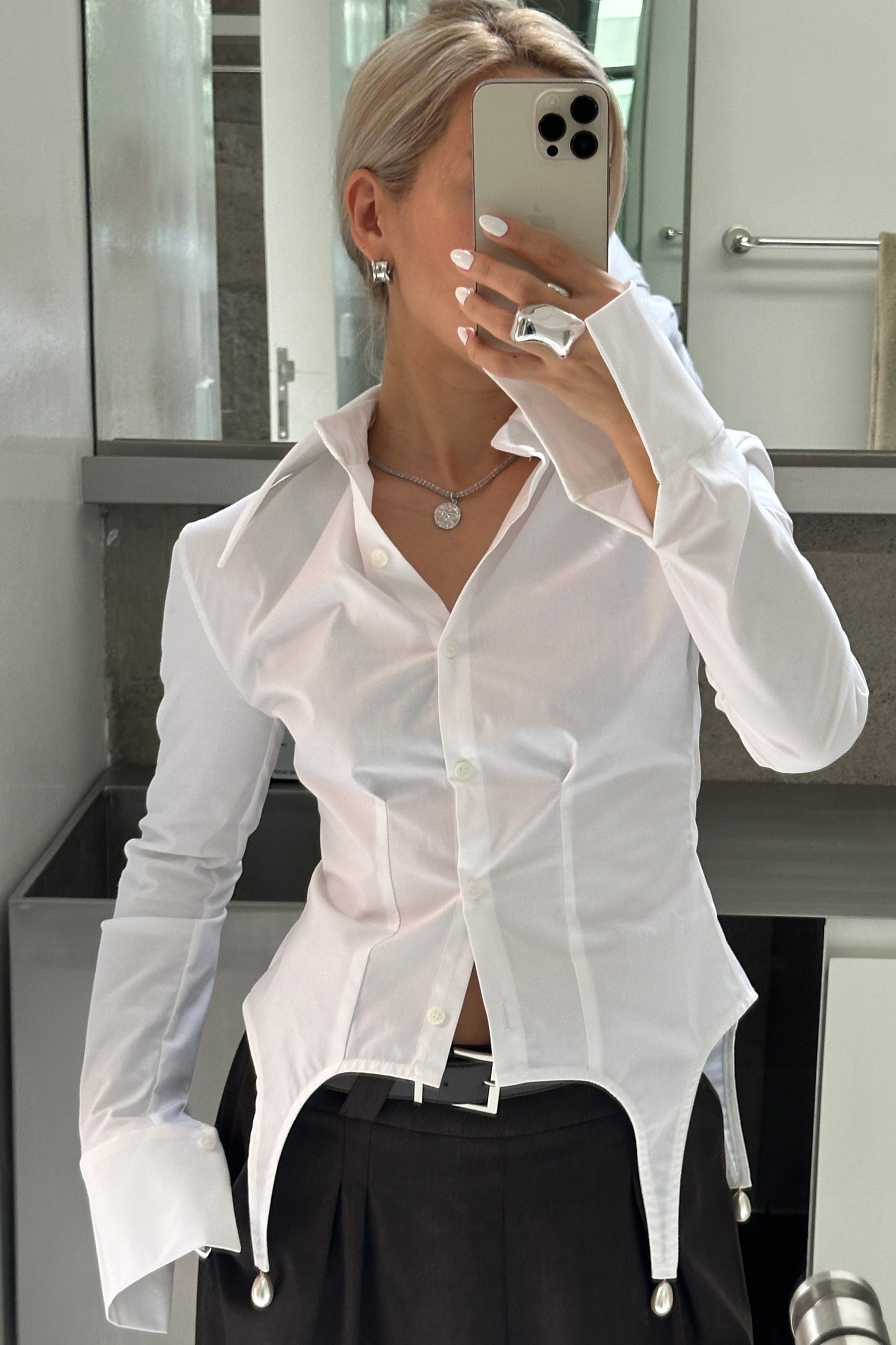 Woman in tailored hip length, long sleeve white button up shirt, with pearl details at the hem and exaggerated collar. Styled half done up and paired with a silver necklace.