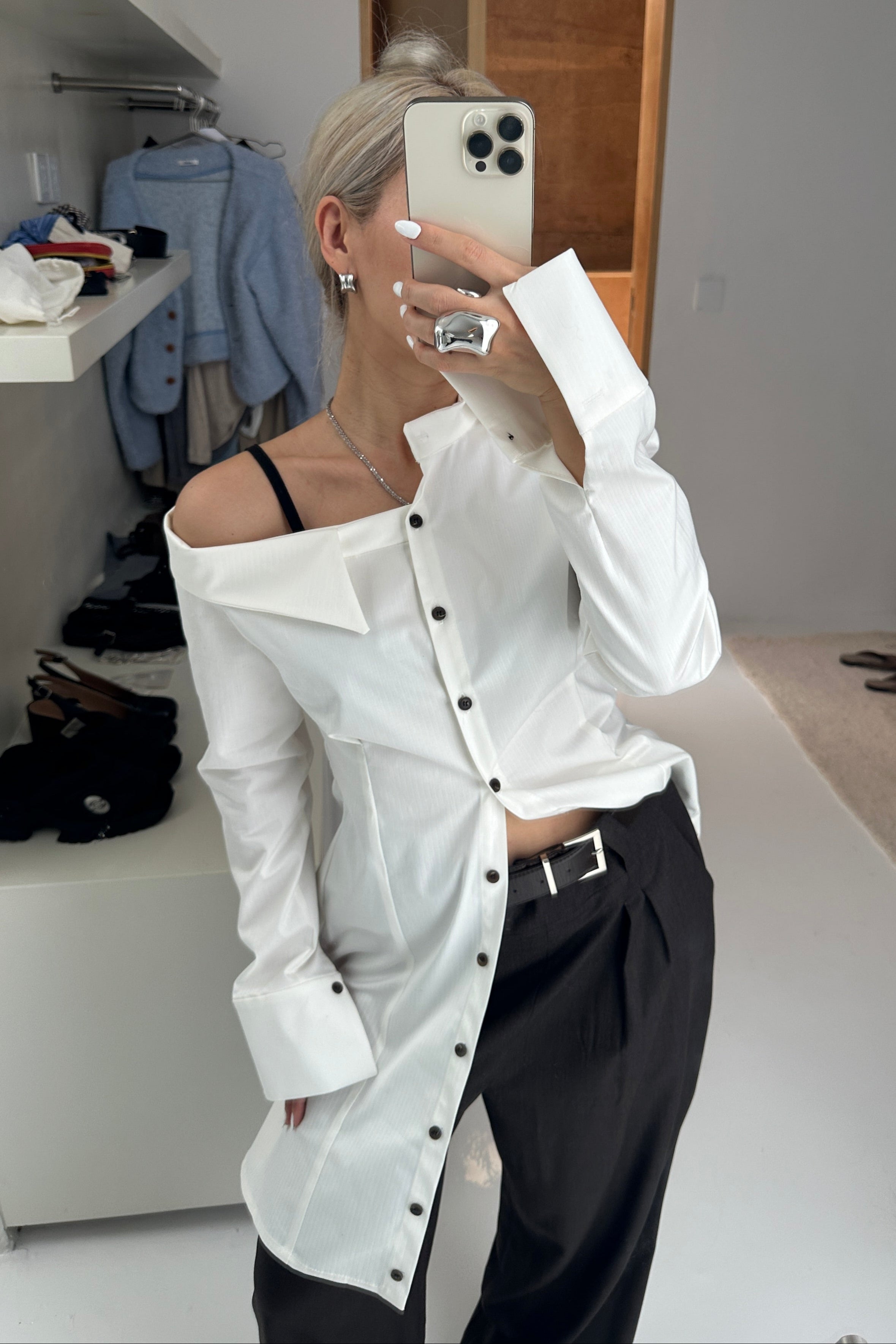 Woman in a tailored white button up shirt dress with asymmetric neckline, off the shoulder detail and black buttons. Styled with black trousers and silver jewellery.