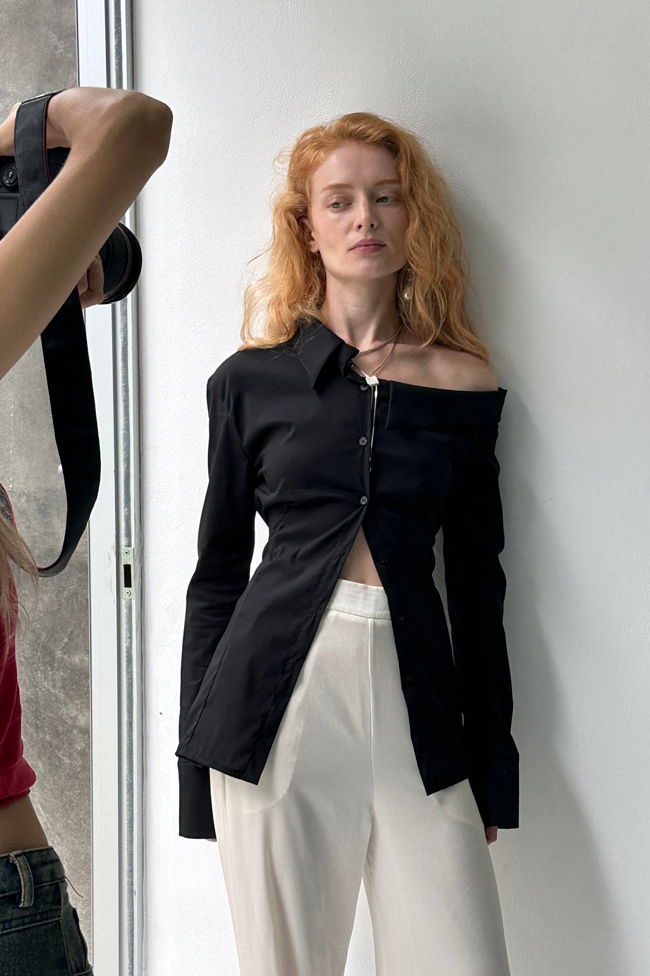 Woman in tailored black button up shirt with asymmetric neckline and off shoulder detail. Styled with cream trousers.