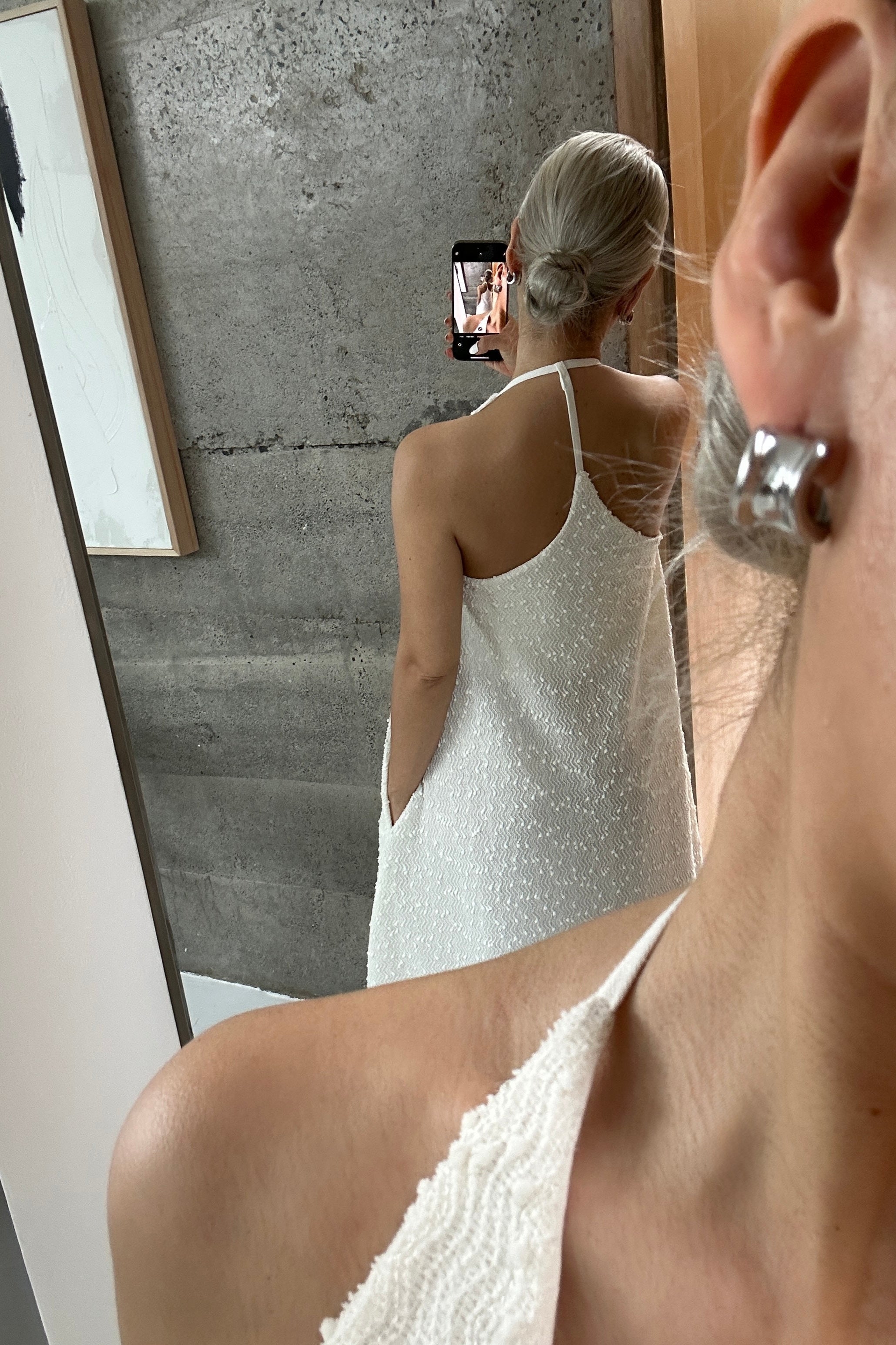 Woman in a white a-line shapeless draped dress with a scooped v-neck and cross back that highlights the shoulders, featuring subtle textural details and fine sequins. Perfect for beach days or Summer outings.