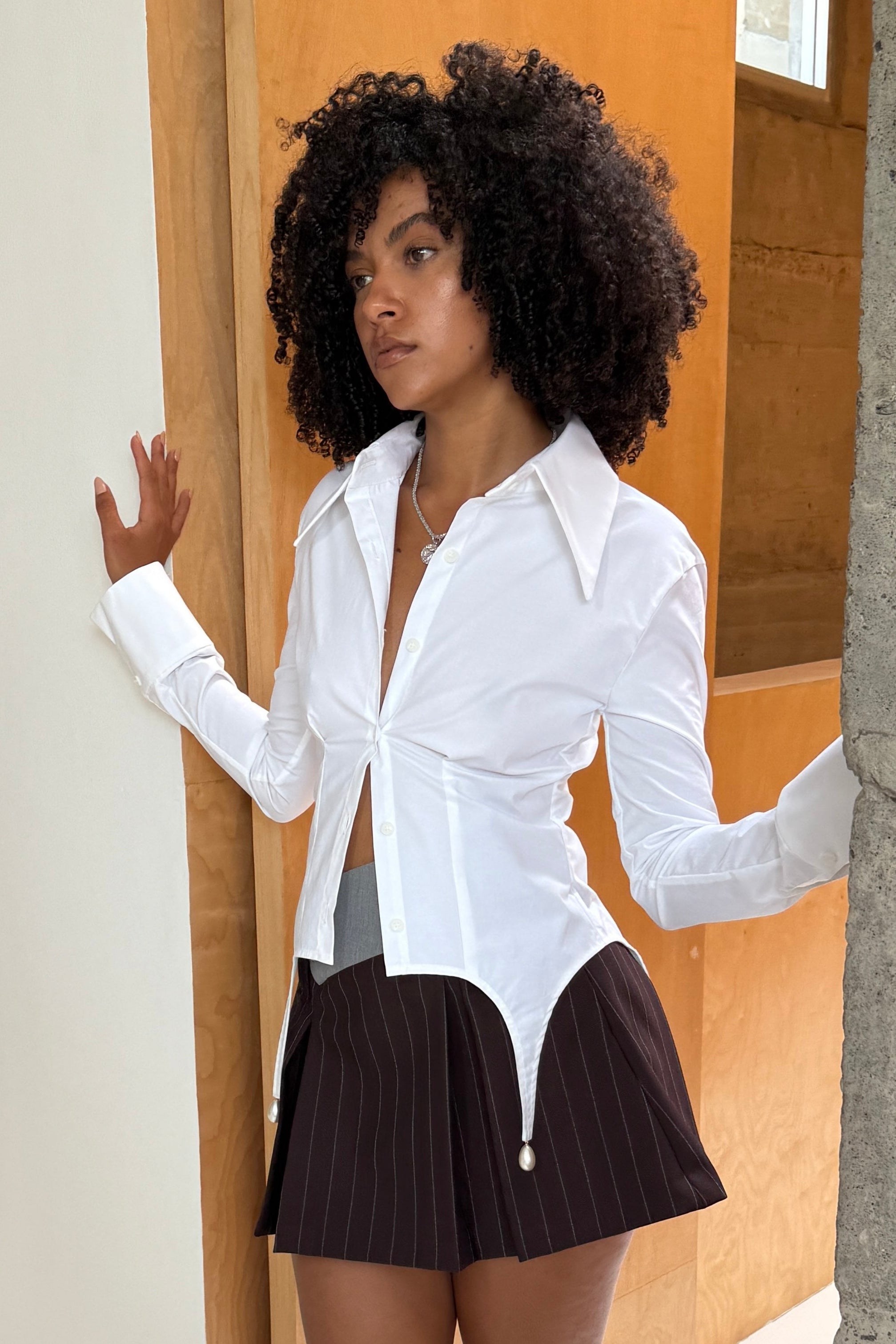 Woman in tailored hip length, long sleeve white button up shirt, with pearl details at the hem and exaggerated collar. Styled with one button done up with a brown mini skirt, paired with a silver necklace.