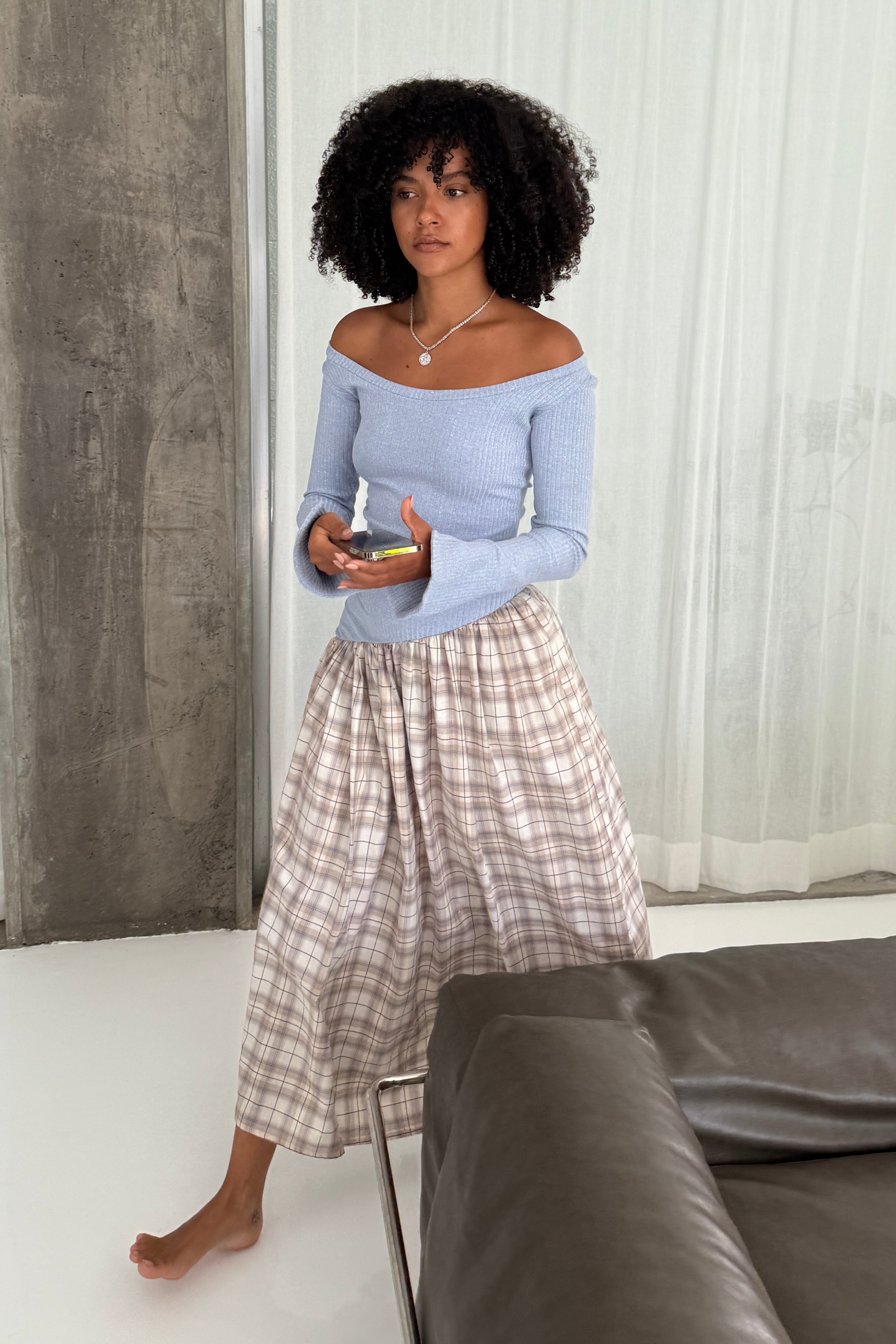 Woman in long sleeve dress, with extra long sleeves, scooped neckline, and artfully gathered asymmetric plaid skirt. Top flatters the figure whilst the skirt flows with every movement. Can be dressed up or down. Styled off shoulder with a silver necklace.
