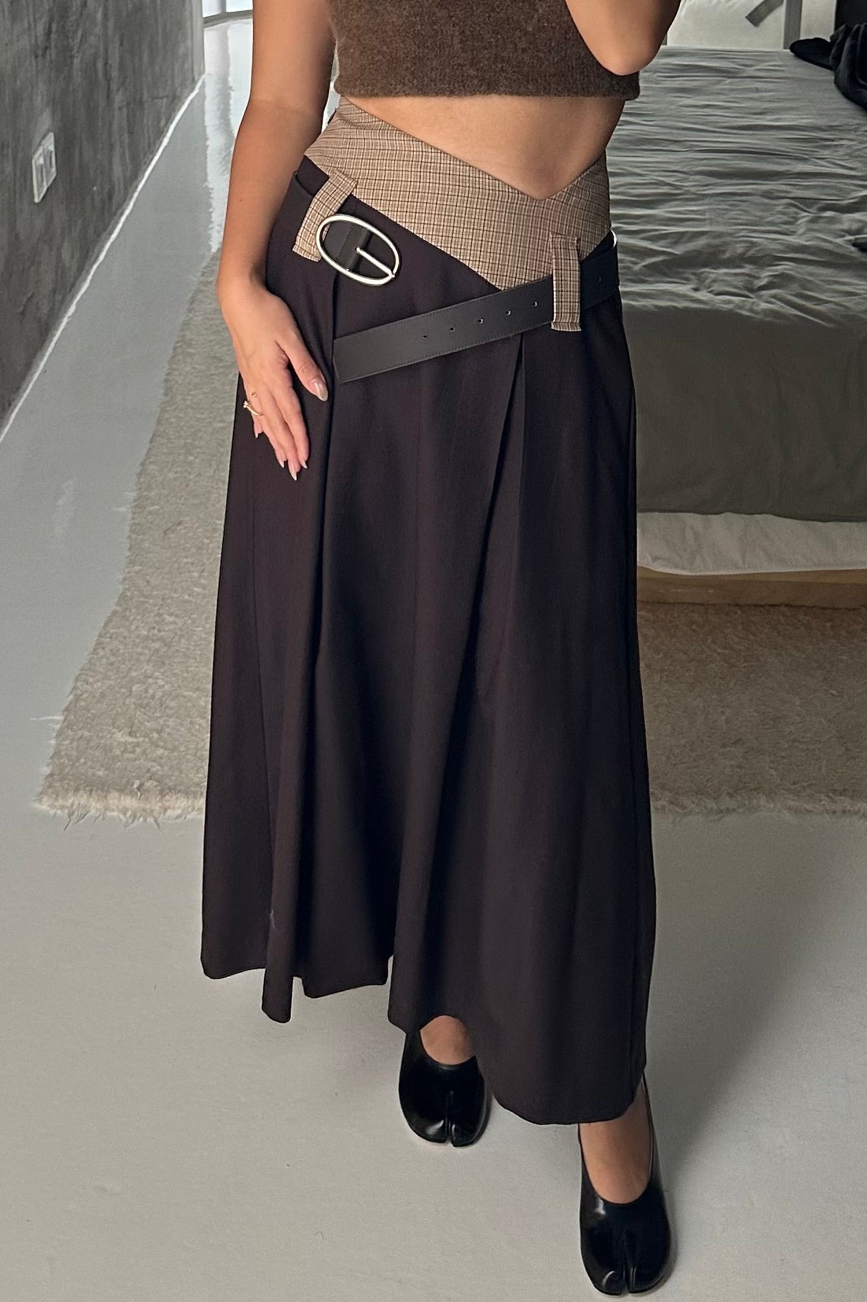 Woman in long skirt with box pleats and dipped waistband. Brown and beige check colourway. Styled with a black belt and tabis.