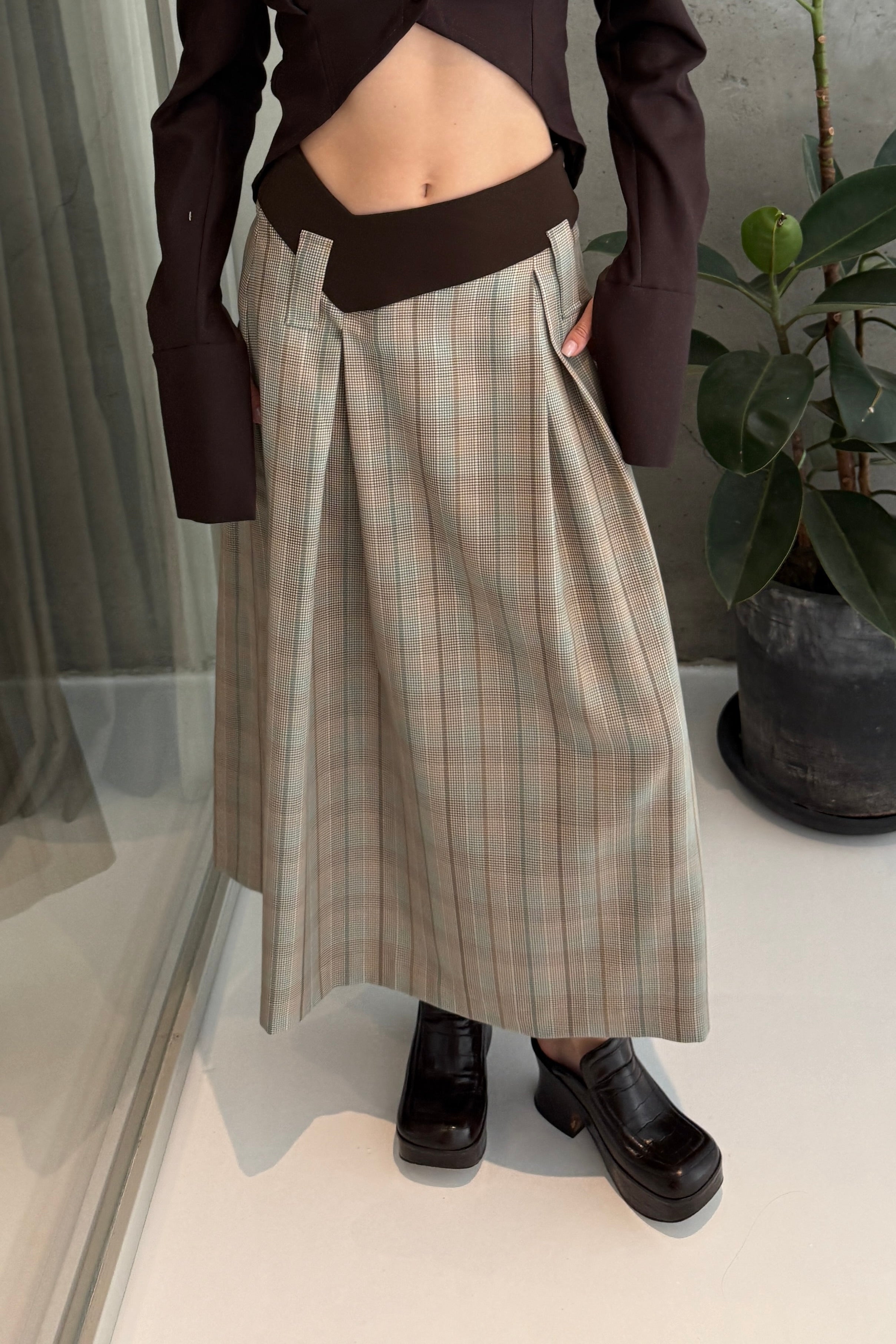 Woman in long skirt with box pleats and dipped waistband. Brown and sage plaid colourway. Styled with a dark brown cropped shirt.