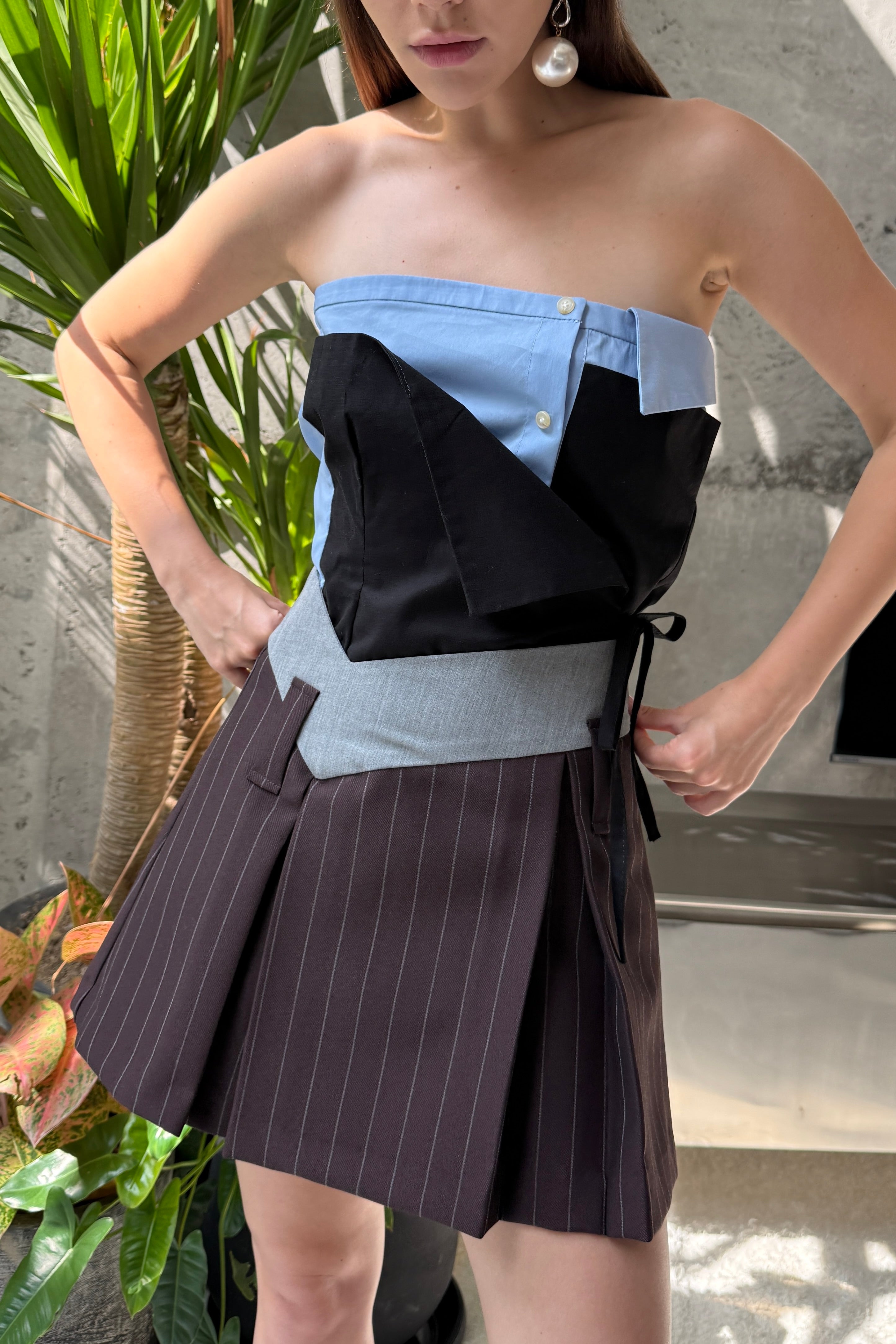 Woman in baby blue strapless mini dress with elements of traditional shirting. Layered black asymmetric tie front detail. Styled as a top, tucked into a brown mini skirt