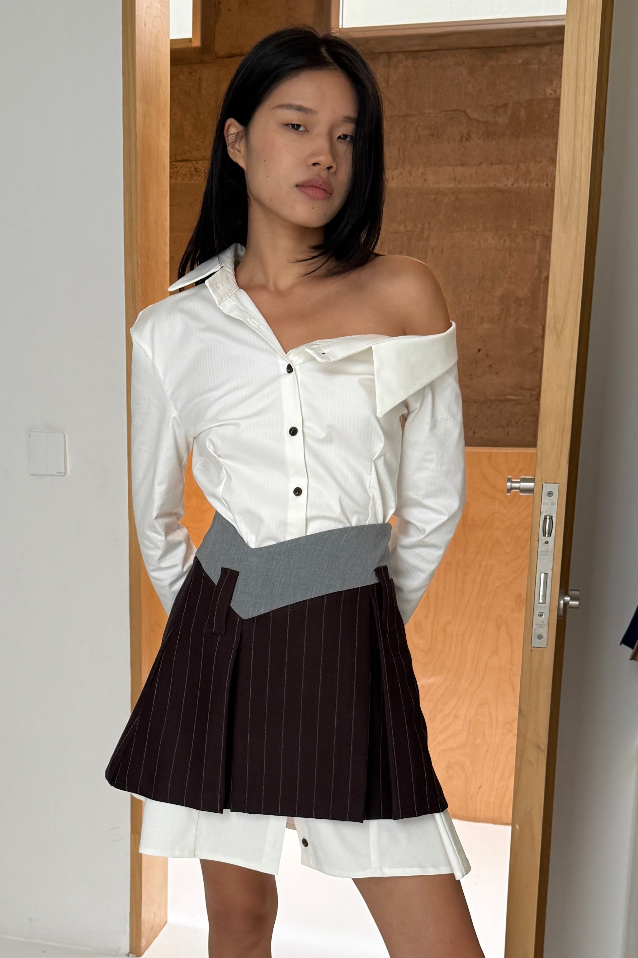 Woman in a tailored white button up shirt dress with asymmetric neckline, off the shoulder detail and black buttons. Layered underneath a brown mini skirt.
