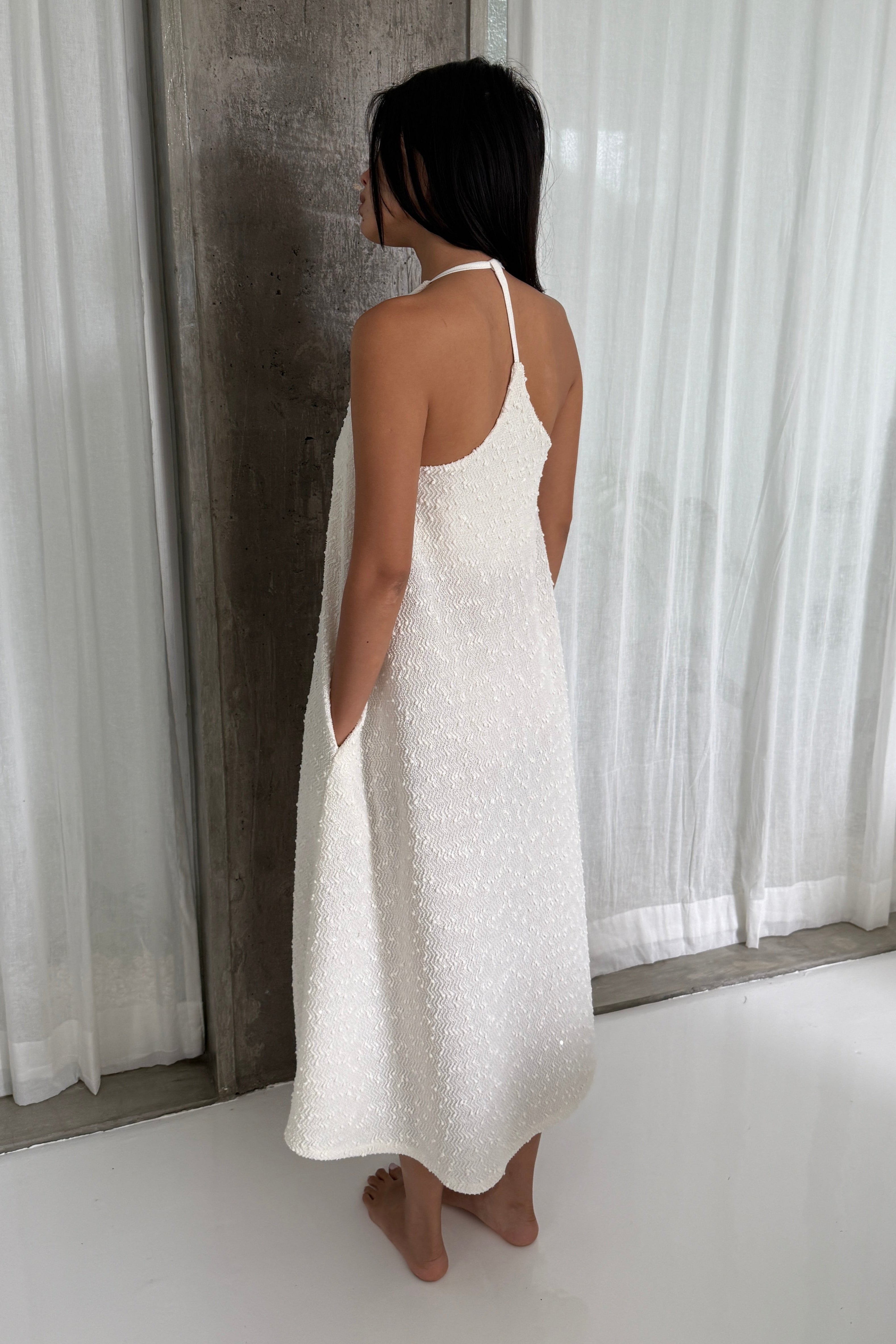 Woman in a white a-line shapeless draped dress with cross back that highlights shoulders, featuring subtle textural details and fine sequins, and hidden side pockets for maximised convenience. Perfect for beach days or Summer outings.