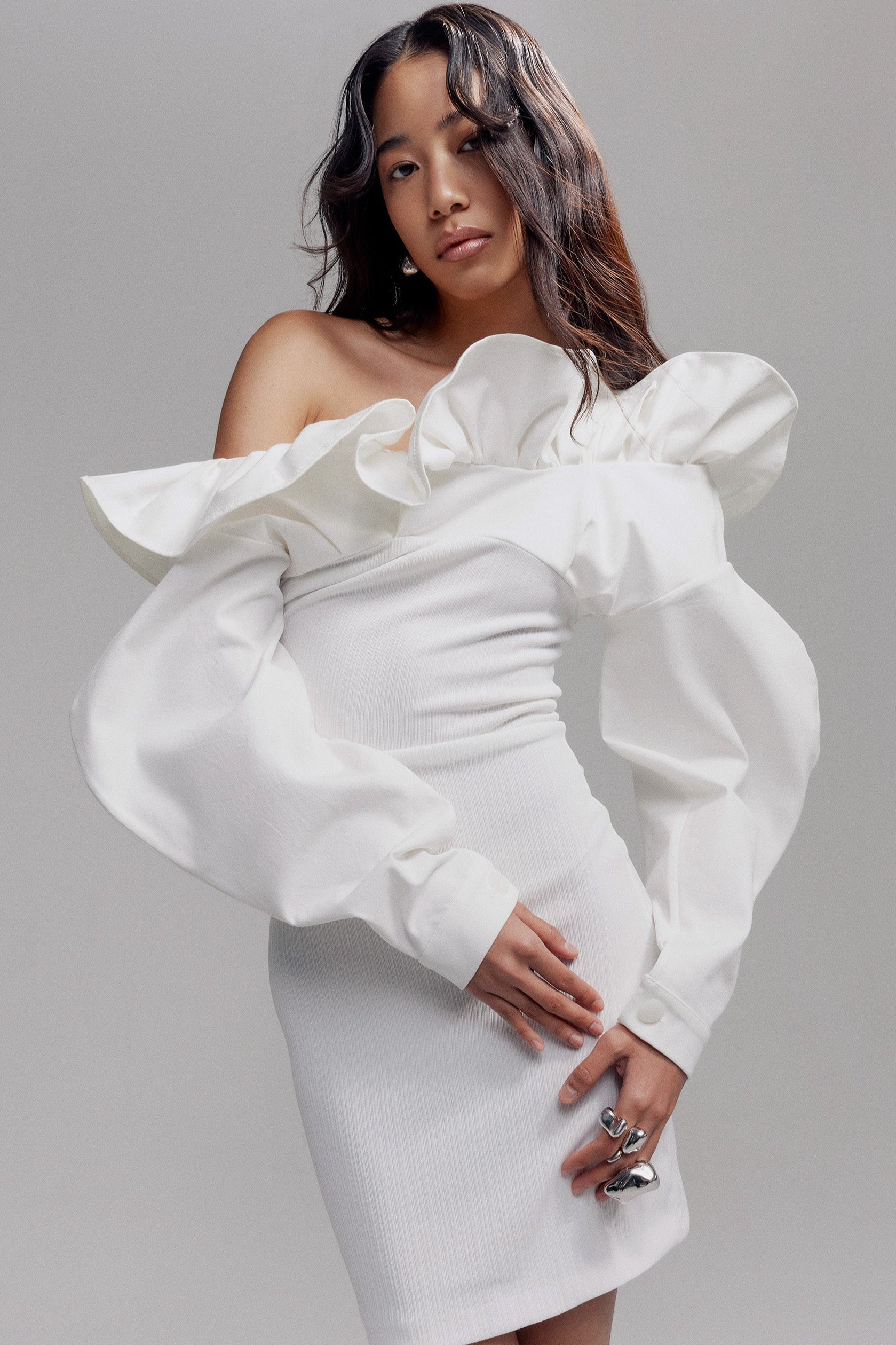 Woman in white off the shoulder, long sleeve mini dress with voluminous sleeves and wave detail around the neck.