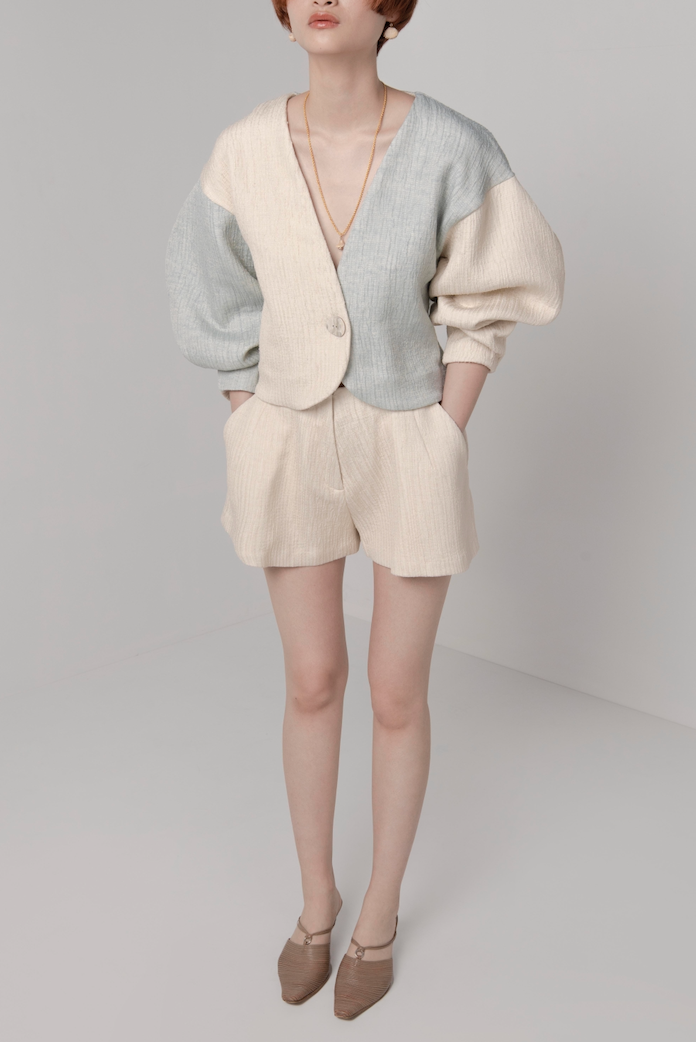 Woman in cream, textured, tailored, high waisted shorts with front pleats, side pockets and fly closure. Styled with matching volume sleeve cardigan.