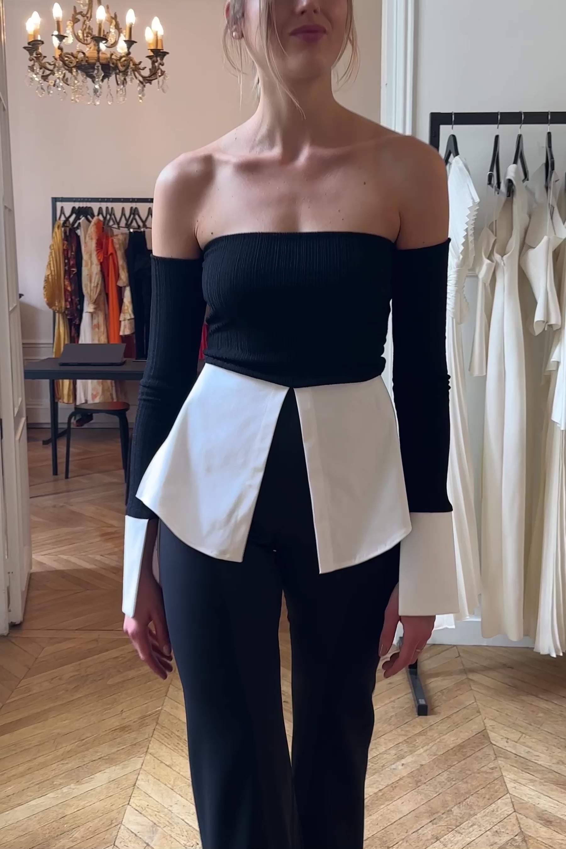 Woman in off shoulder black crop top with white shirting details on the cuffs and hem. Styled with black trousers.