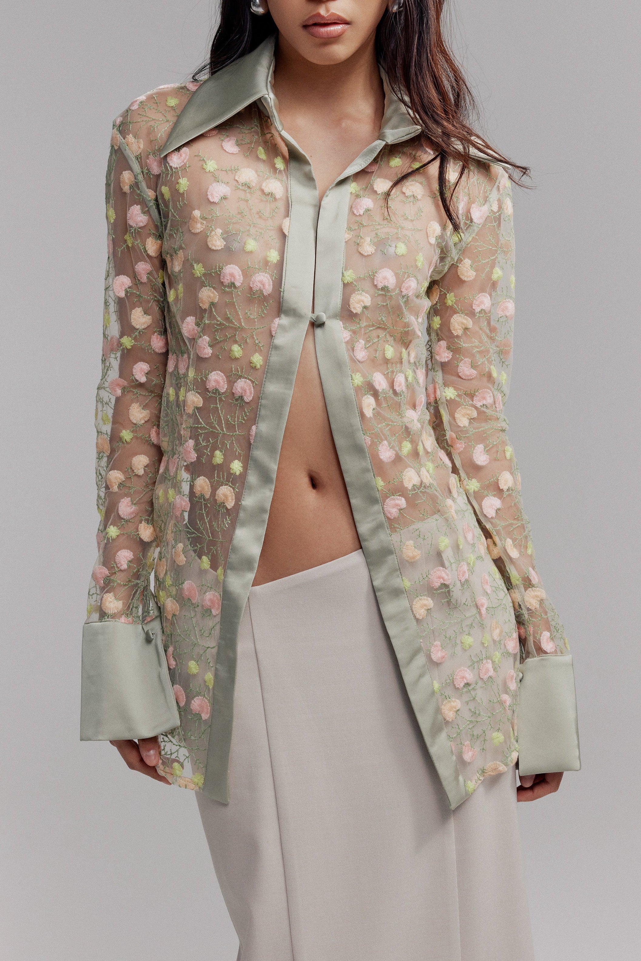 Woman in sage green sheer button down shirt. Featuring opaque collar and cuffs and pink and green floral embroidered detail. Styled with a cream maxi skirt.
