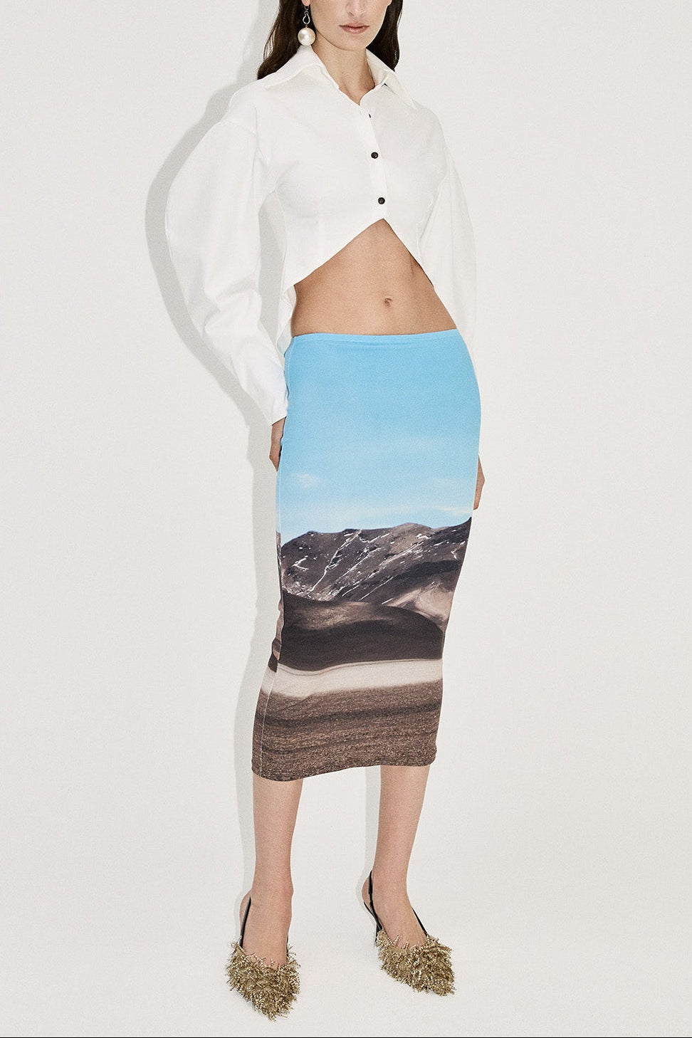 Woman in volume sleeve white button up shirt with cornered hem. Styled with a landscape print pencil skirt.
