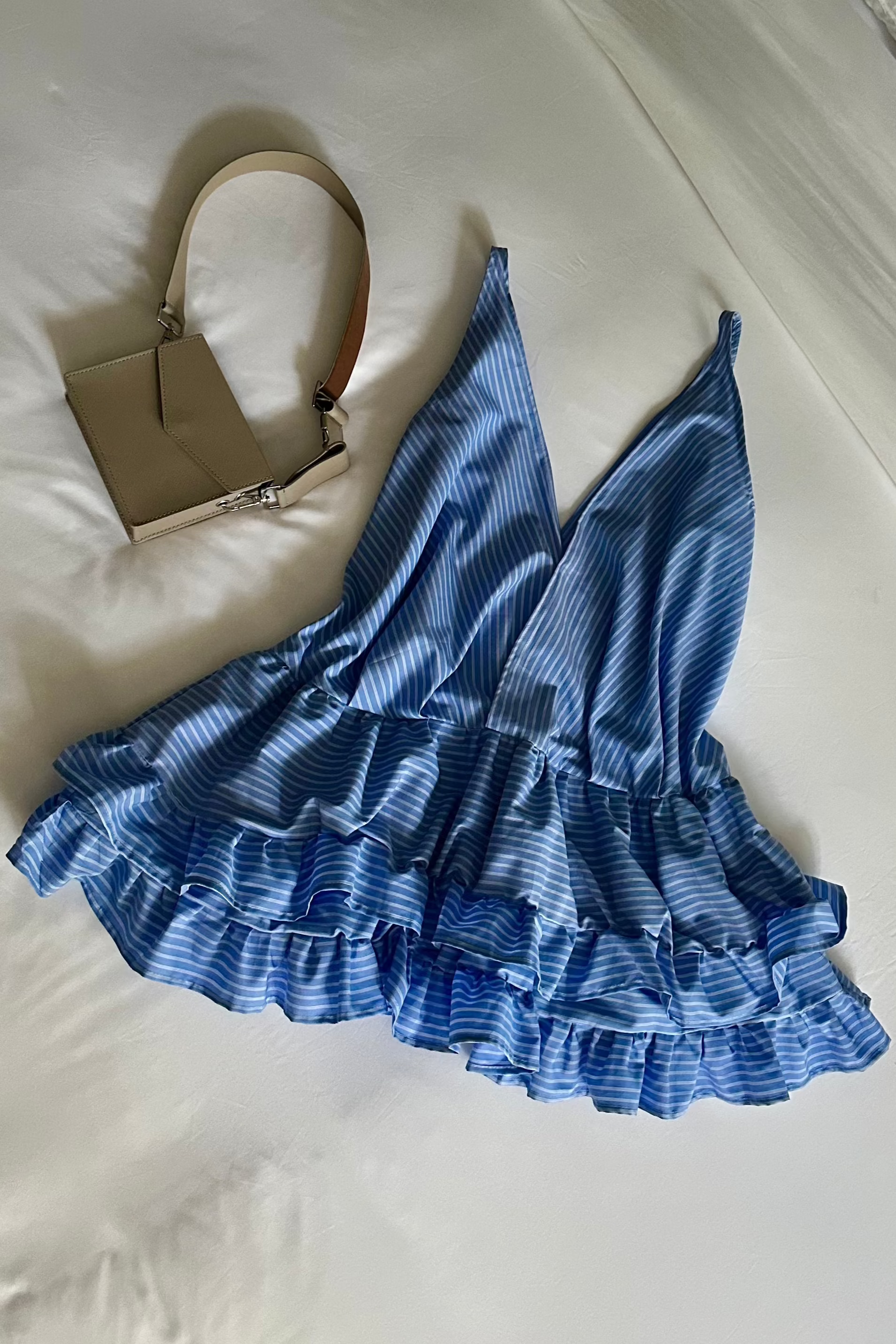 Flat lay of baby blue striped deep v-neck mini ruffled dress. The perfect beach cover up or casual summer dress. Paired with cream bag.