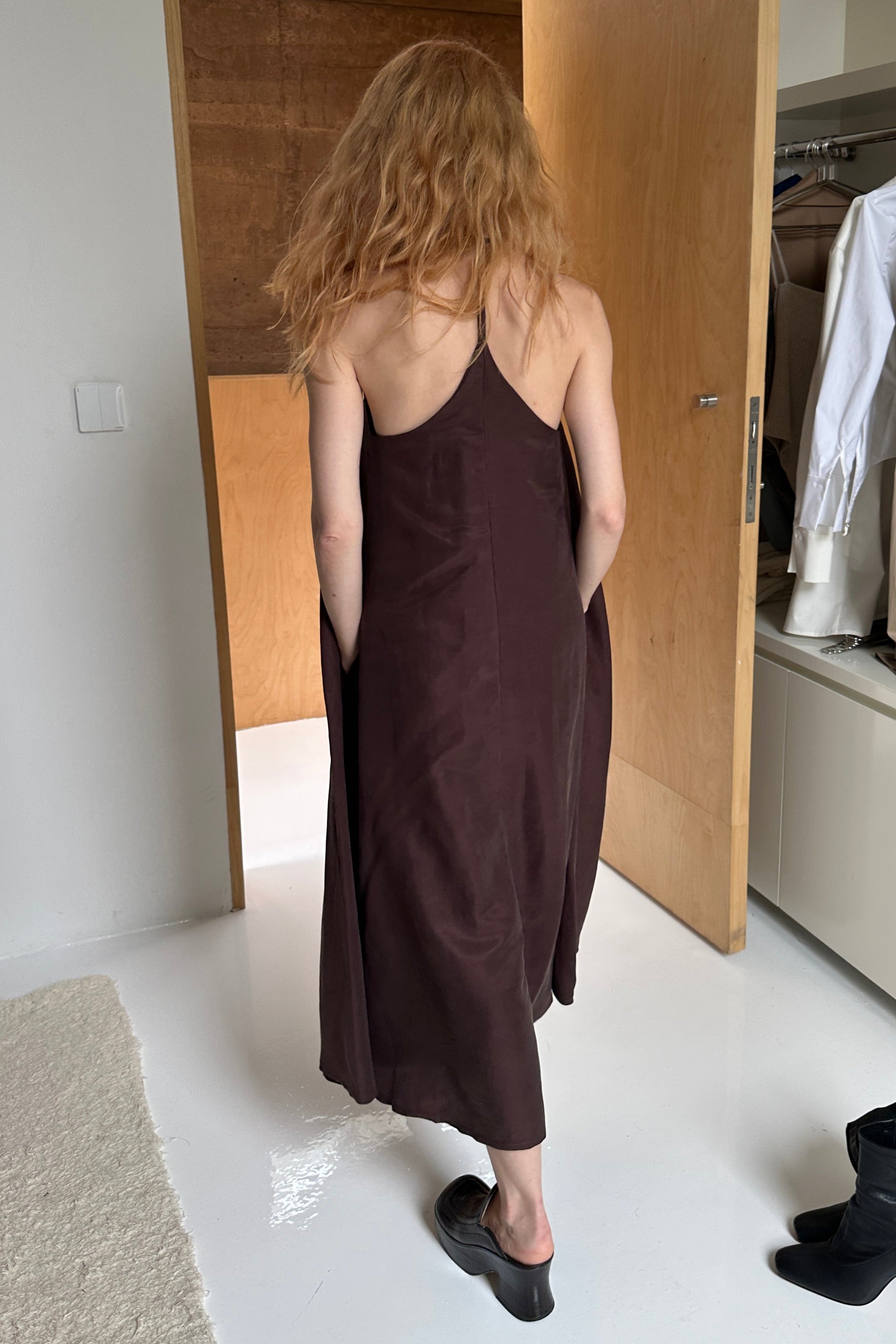 Woman in a brown a-line shapeless draped dress with crossed, open shoulder back. 