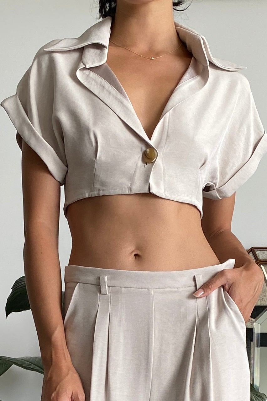 Woman in cream tailored crop shirt with short sleeves and singular button front closure. Featuring front darts and dramatic collar. Styled with matching wide leg pants.