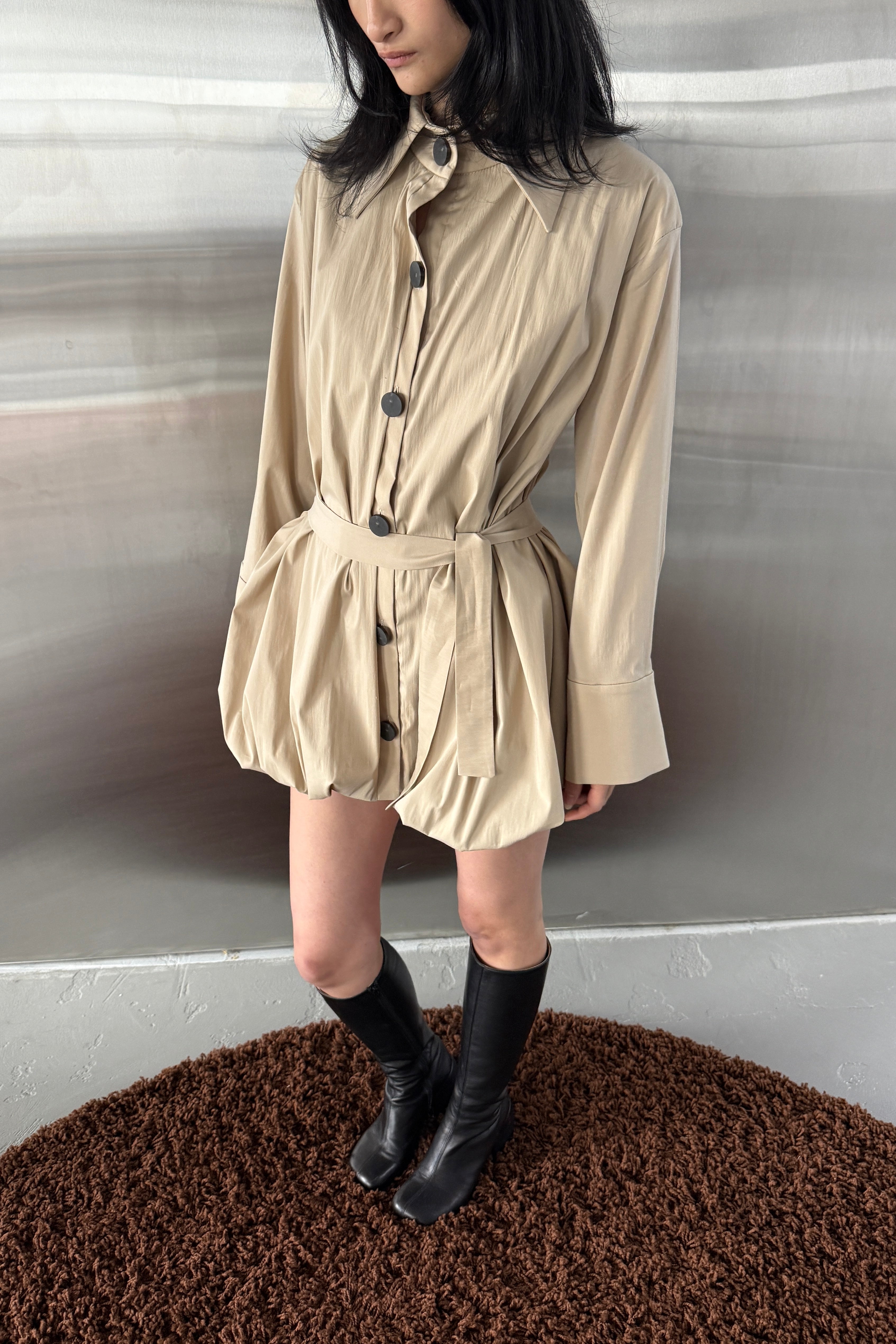 Woman in beige balloon hem jacket dress. Black button front closure, cinched with detachable tie. Styled with black boots.