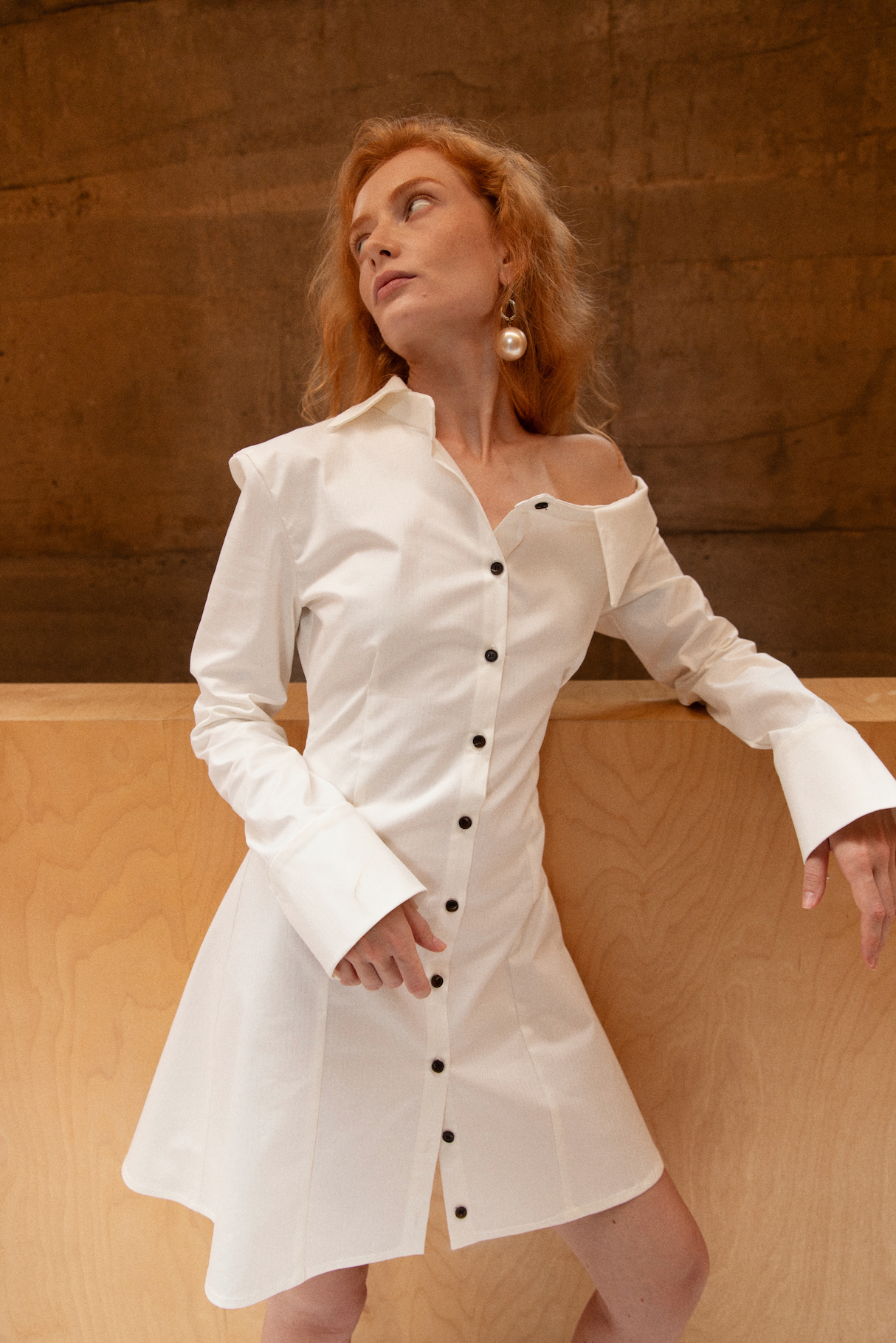Woman in a tailored white button up shirt dress with asymmetric neckline, off the shoulder detail and black buttons. Styled with large pearl earring.