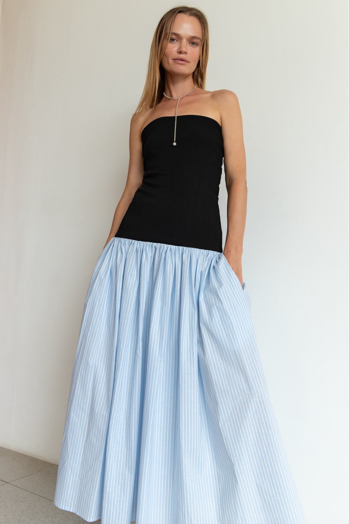 Woman wearing a black strapless top and light blue striped skirt against a white wall.