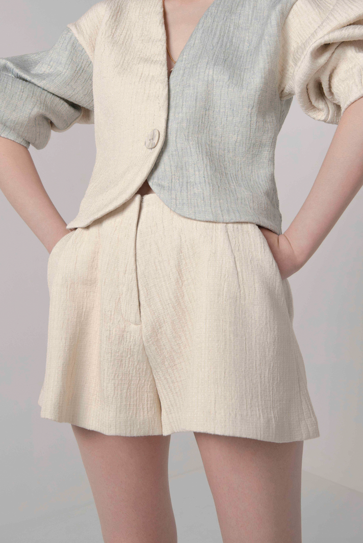Woman in cream, textured, tailored, high waisted shorts with front pleats, side pockets and fly closure. Styled with matching volume sleeve cardigan.