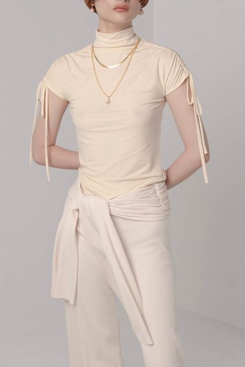 Woman in cream turtle neck knit t-shirt with ruching ties on the sleeves. Styled with matching cream pants with tie detail and gold jewellery.