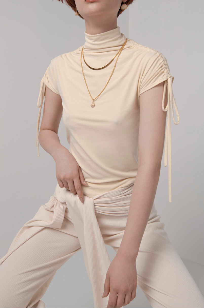 Woman in cream turtle neck knit t-shirt with ruching ties on the sleeves. Styled with matching cream pants with tie detail and gold jewellery.